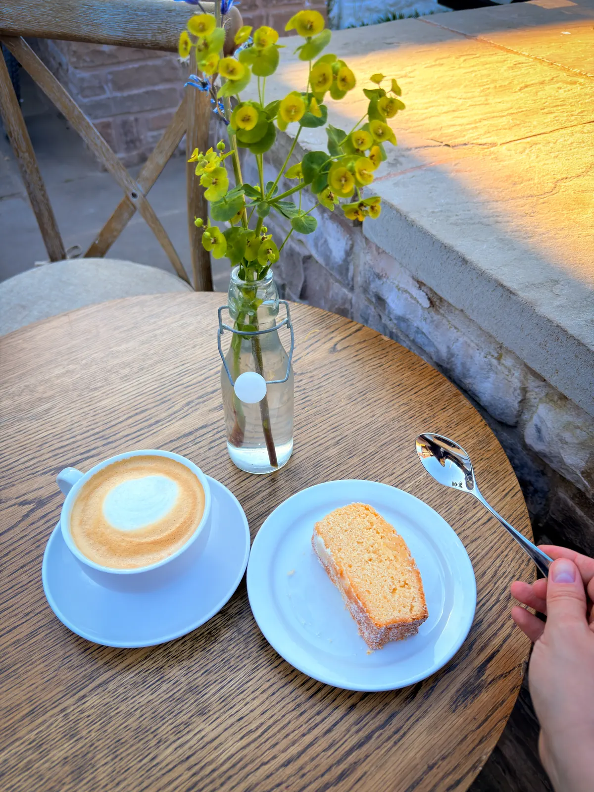 A flat white and a slice of lemon drizzle cake on an oak table in the sun, with a jar of yellow flowers.